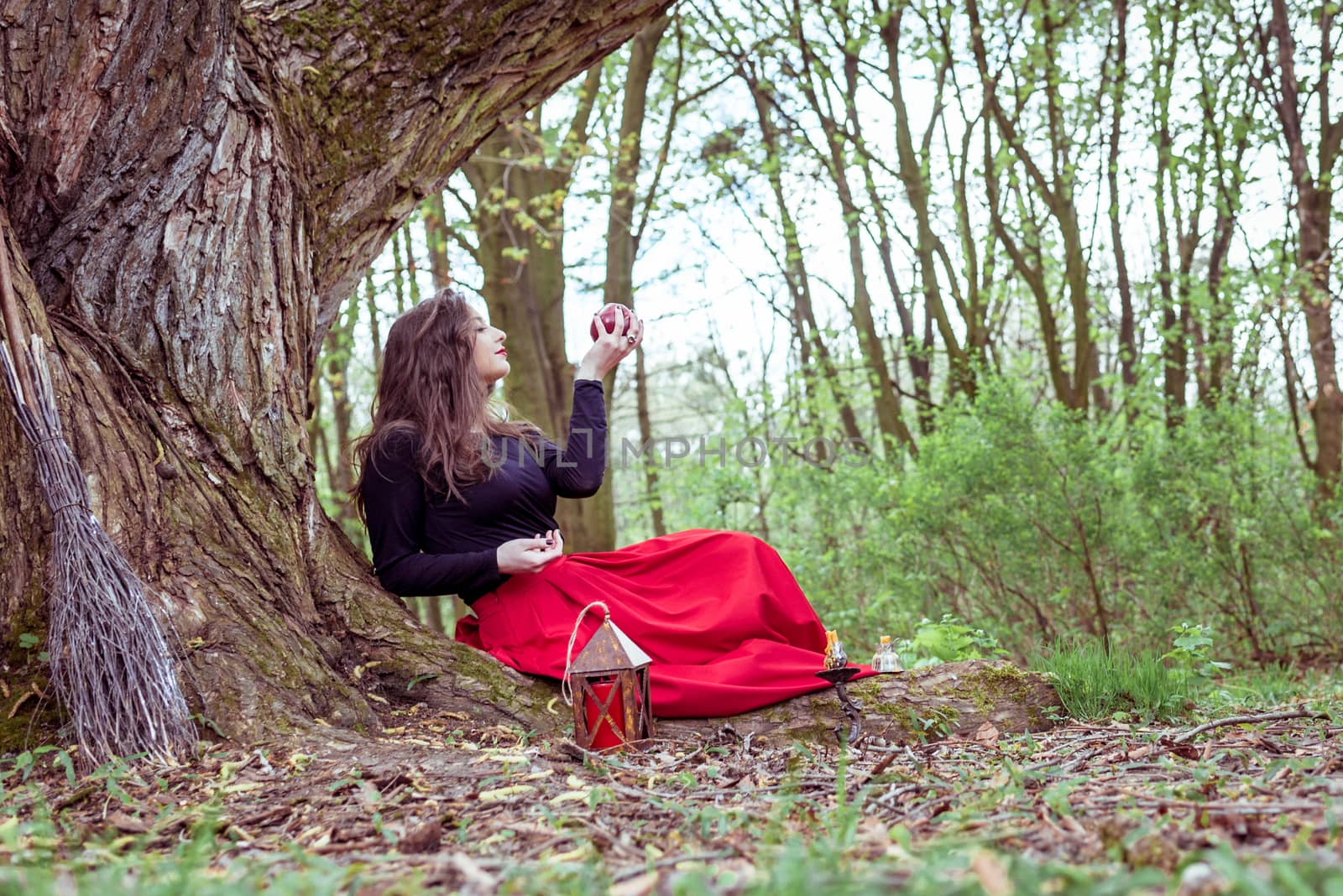 mystical witch woman in red dress sitting under a tree and holding an apple in the park