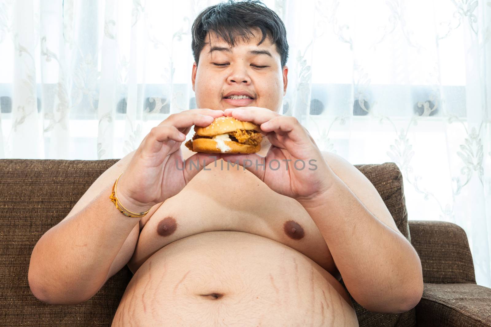 Asian fat man eating hamburger seated on armchair