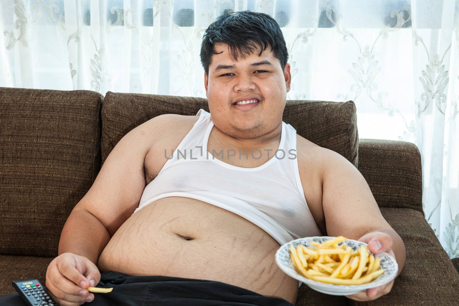 Lazy overweight asian male sitting with fast food on couch and watching television