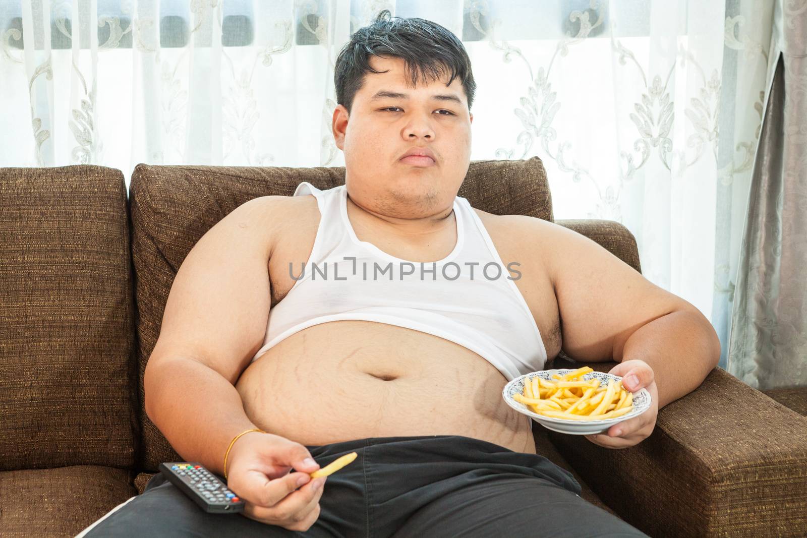 Lazy overweight asian male sitting with fast food on couch and watching television