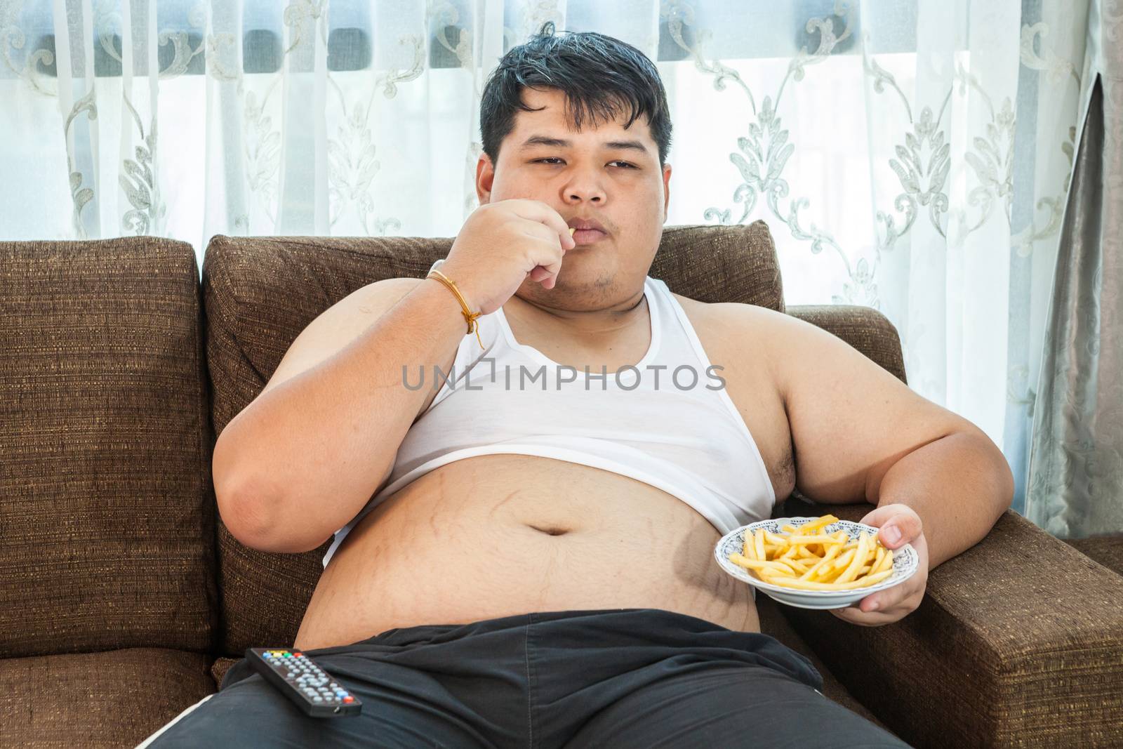 Lazy overweight asian male sitting with fast food on couch and watching television