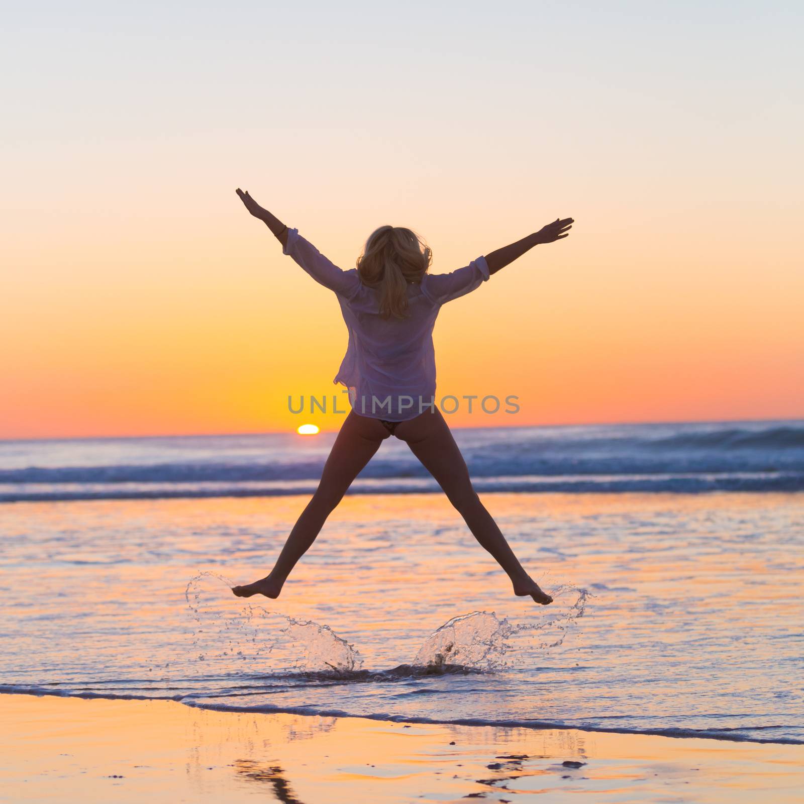 Young beautiful woman wearing jeans shorts and white t-shirt jumping on Cofete beach, Fuerteventura, Spain in sunset.