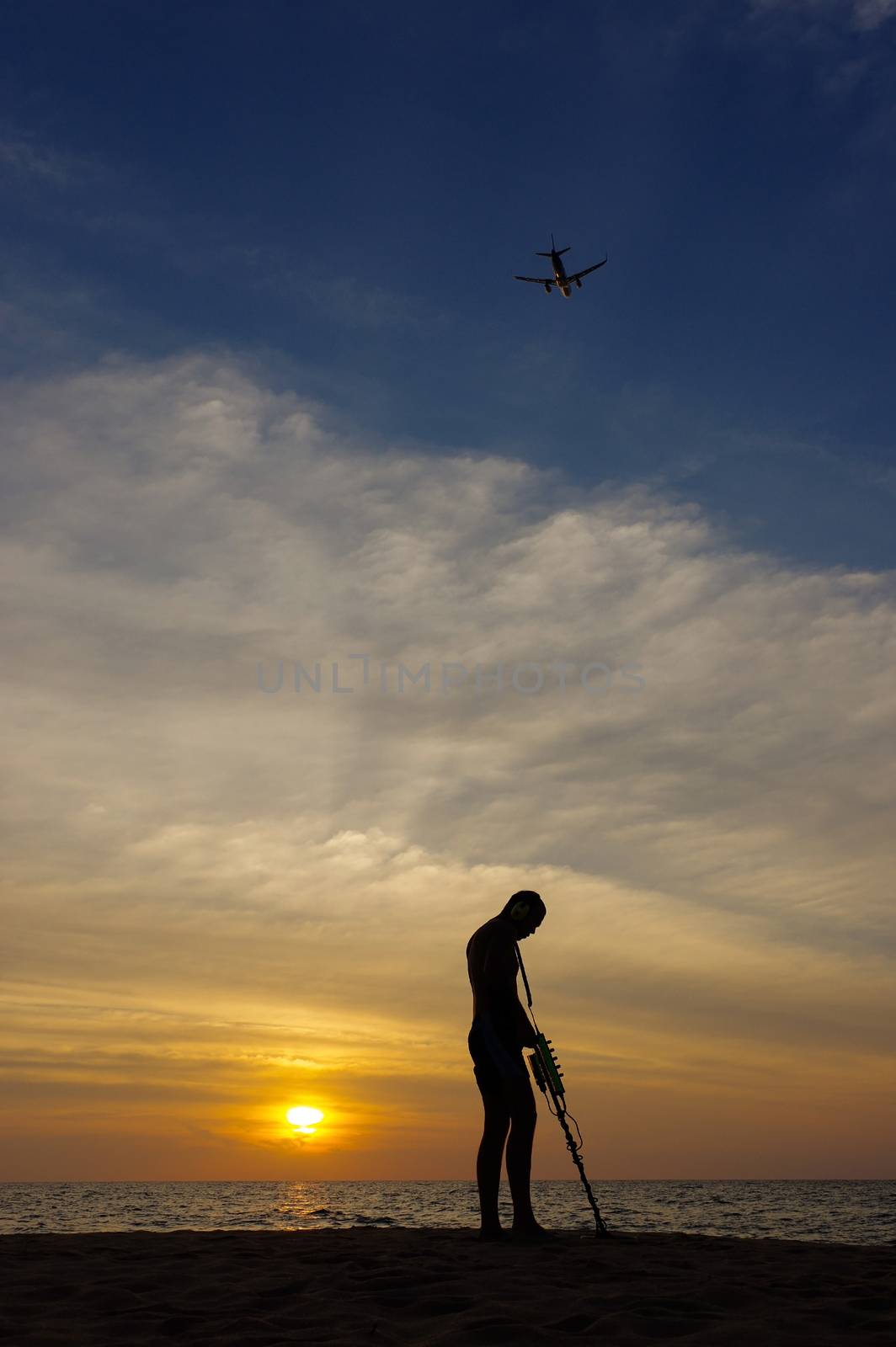 a treasure hunter with Metal detector on sunset on the beach with a plan in the sky