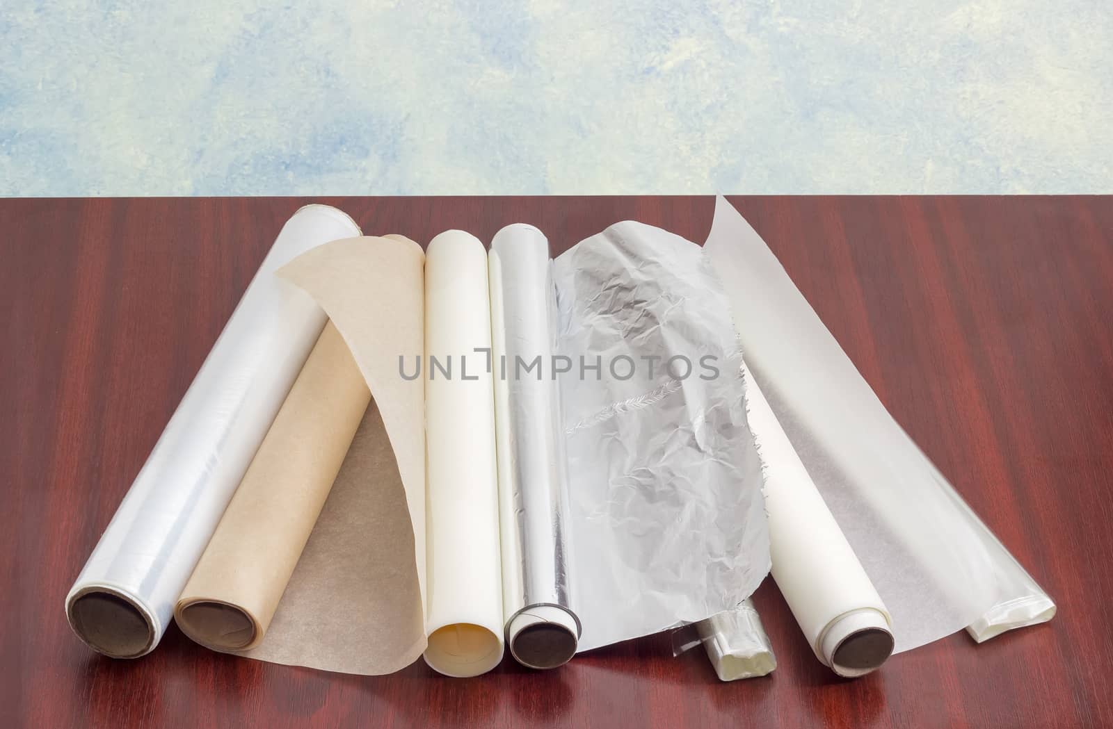 Several rolls of the plastic oven bags, plastic food wrap, aluminum foil and various parchment paper for household use on a dark red wooden surface
