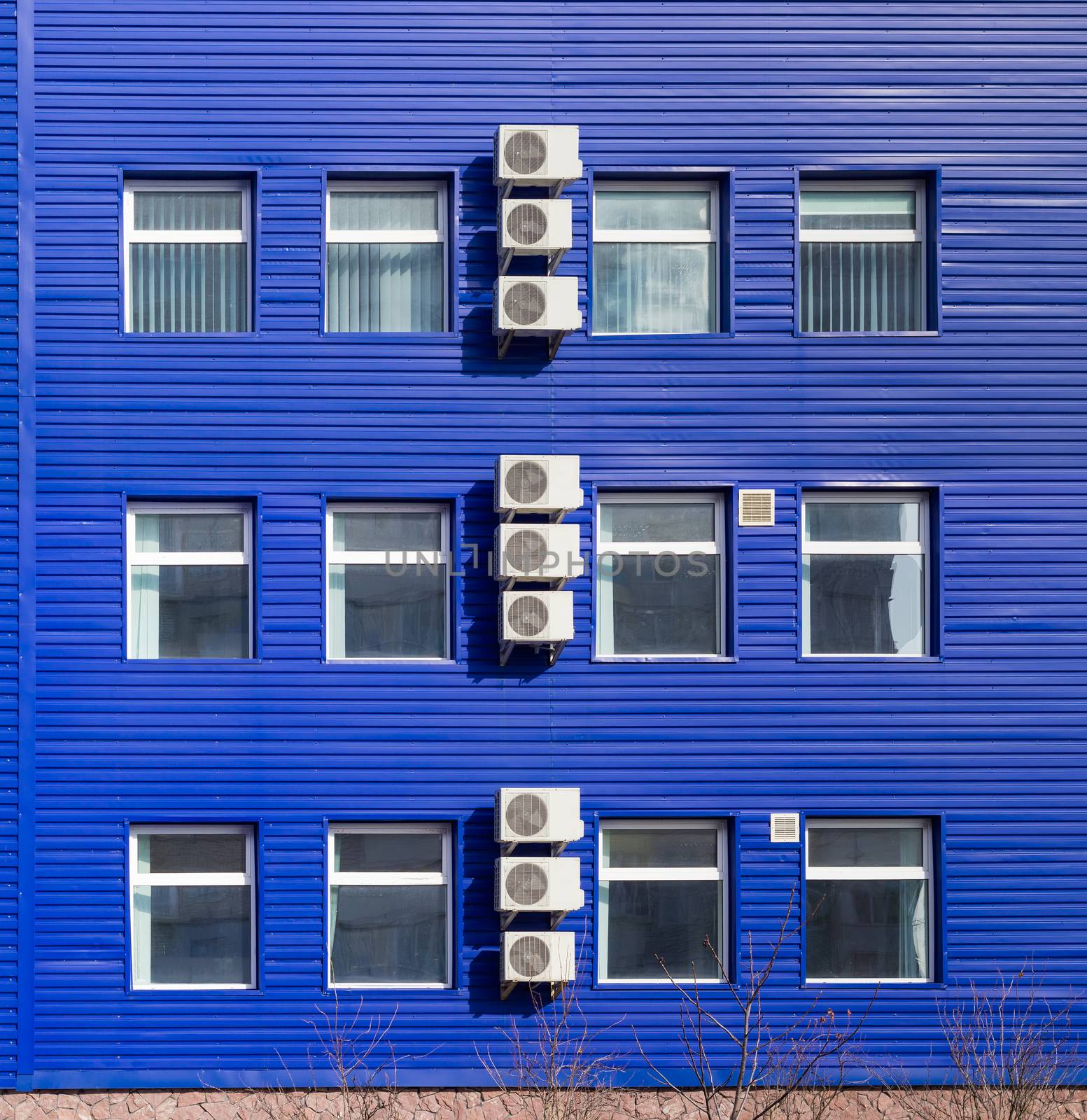 Fragment of the blue wall of a small office building with several window and vertical row of the outside parts of a split-type air conditioners
