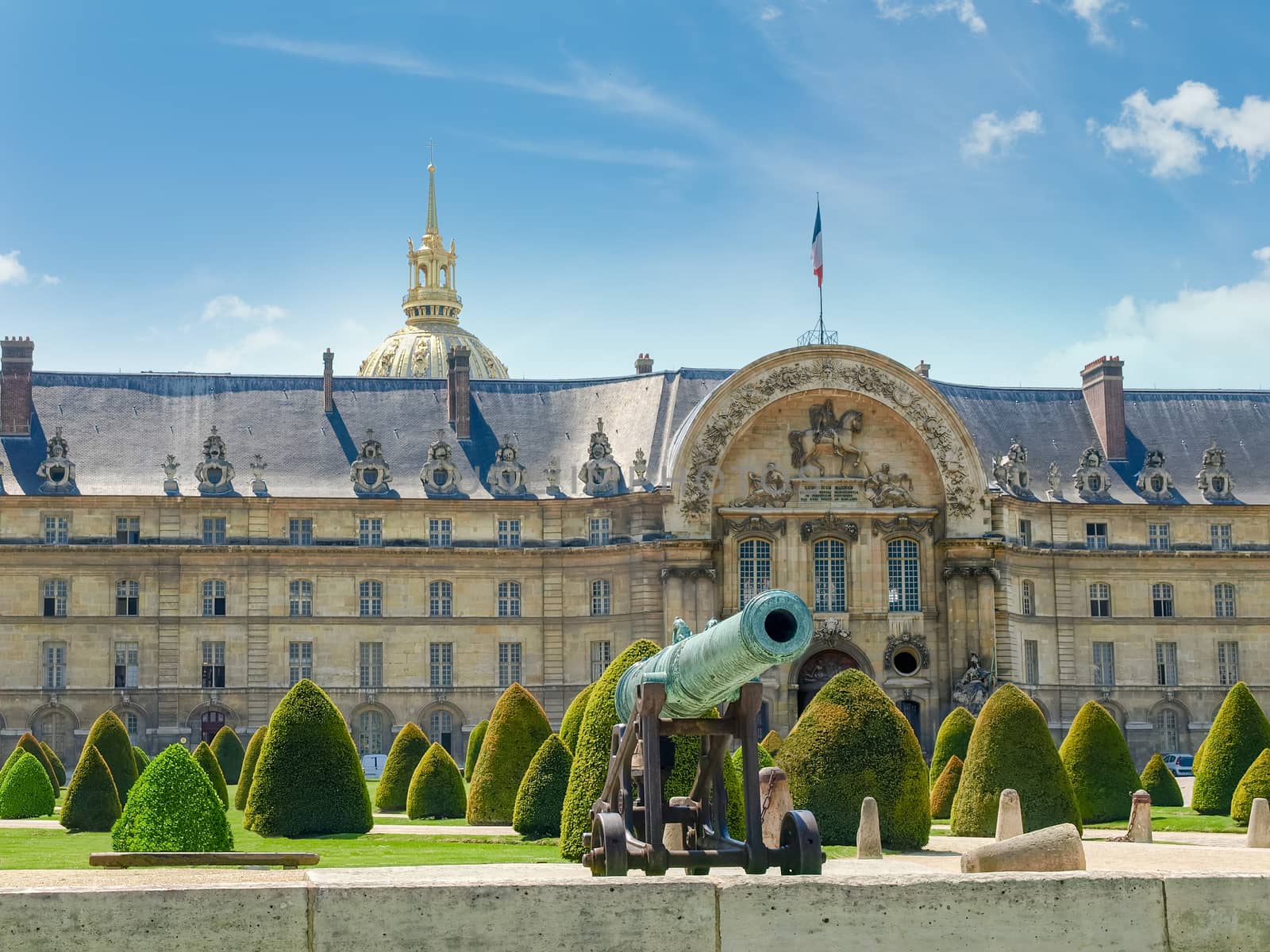 Part of the northern facade of the Hotel Des Invalides with archway of a main entrance with low-relief of King of France Louis XIV, bronze cannon and garden in the foreground in Paris
