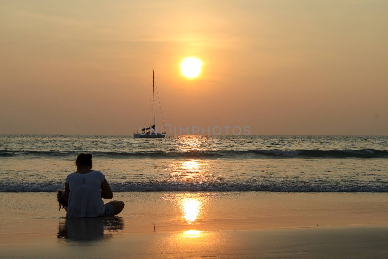 A girl sitting by the sea at sunset and looking at a passing boat, in a light summer dress. She looks at the sea. Sun sets over the horizon.