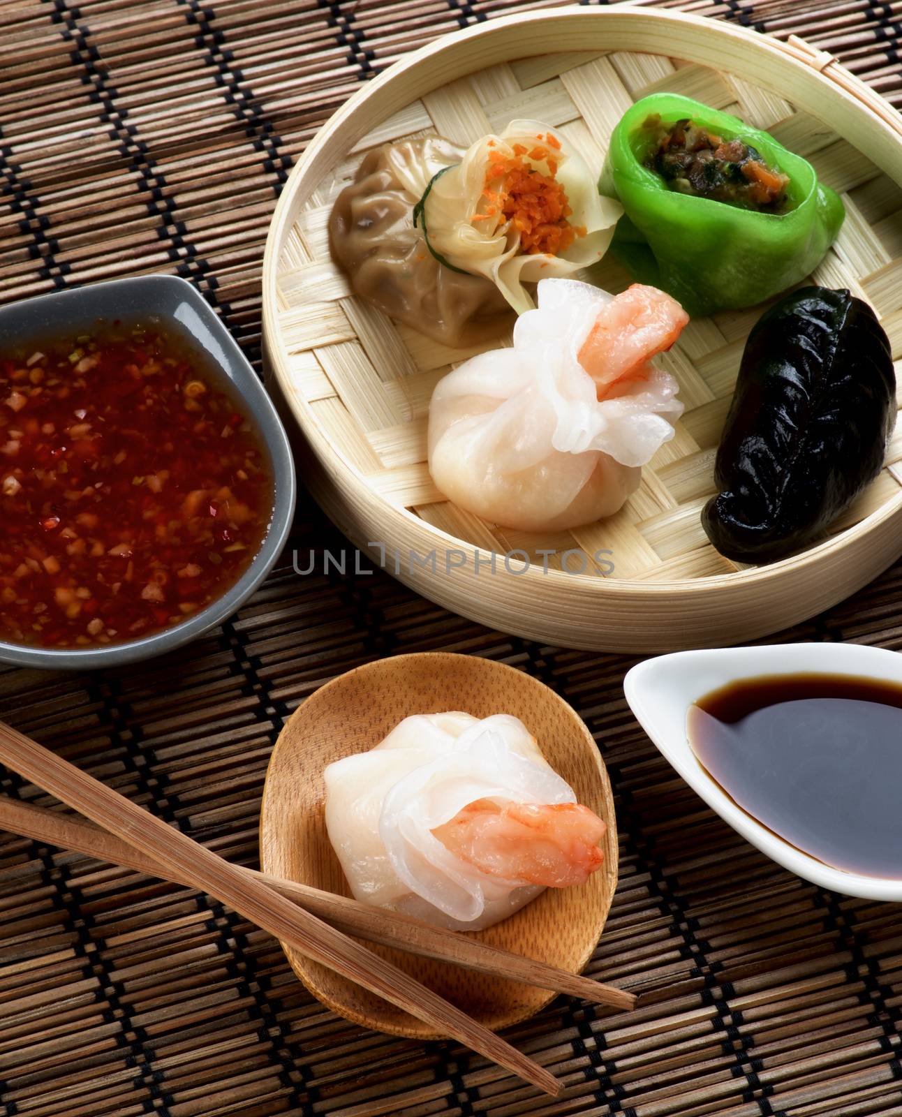 Assorted Dim Sum in Bamboo Steamed Bowl and Tori with Shrimp on Wooden Plate with Red Chili and Soy Sauces and Chopsticks closeup on Straw Mat background