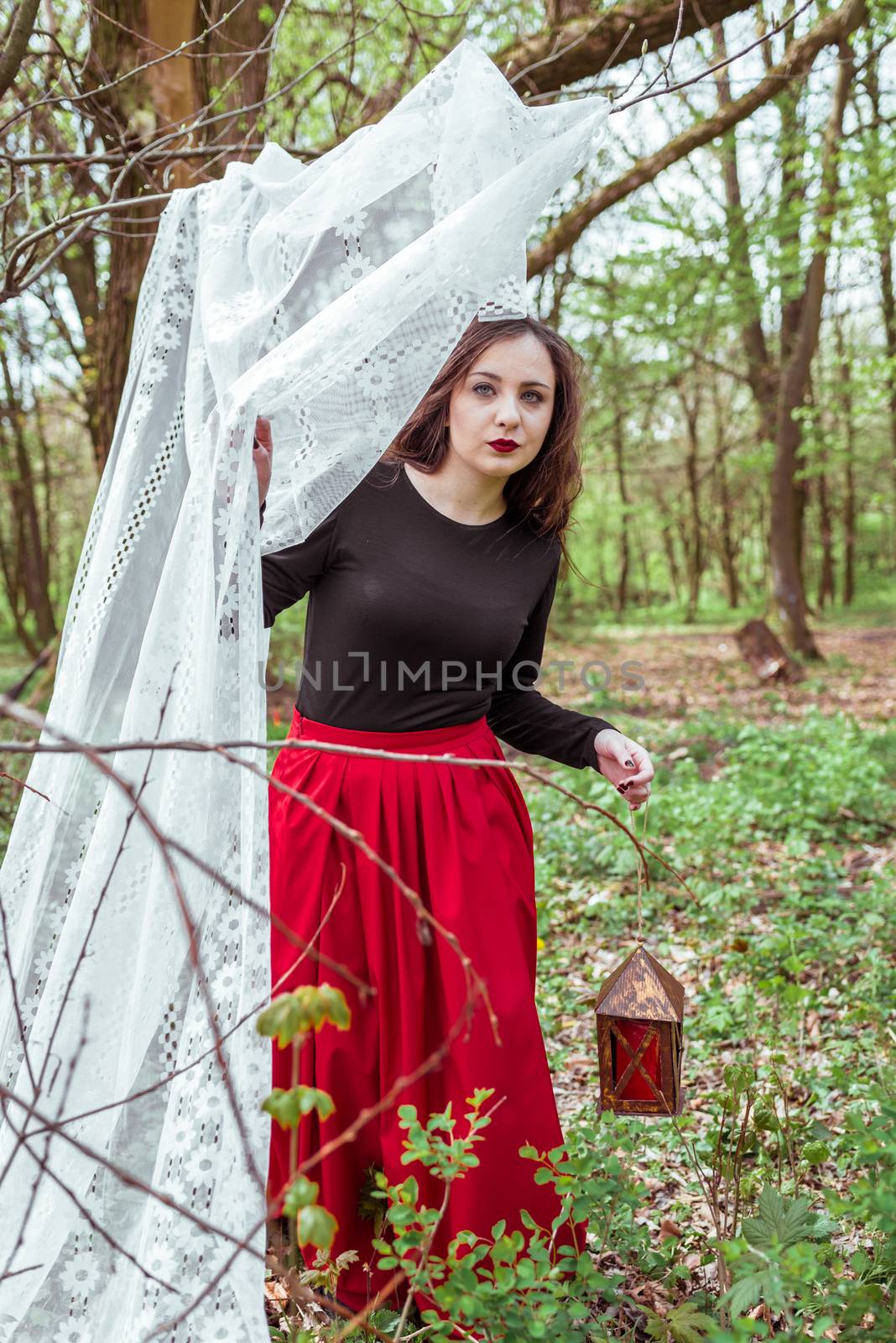 mystical witch woman in a red skirt with a lantern hidden behind the curtains in spring forest