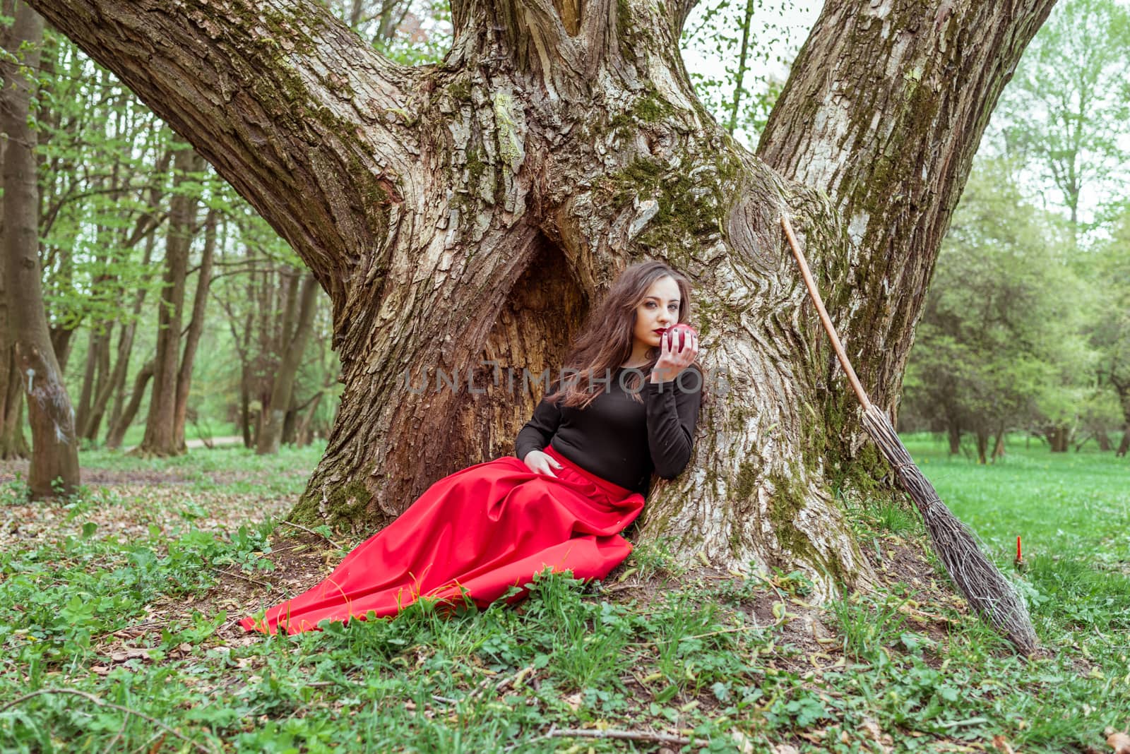 mystical witch woman in red dress sitting under a tree and holding an apple in the spring forest