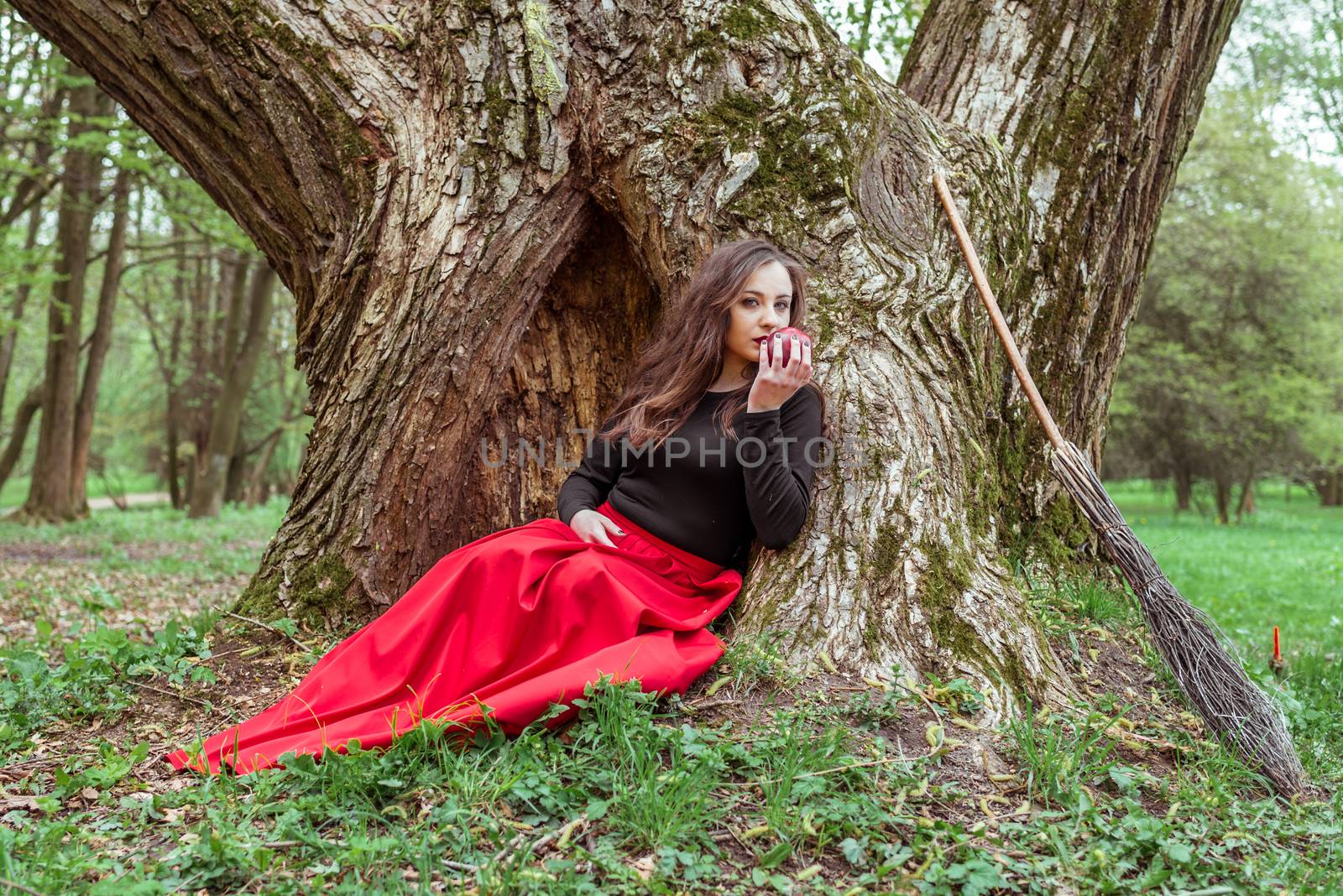 mystical witch woman in red dress sitting under a old tree in the forest