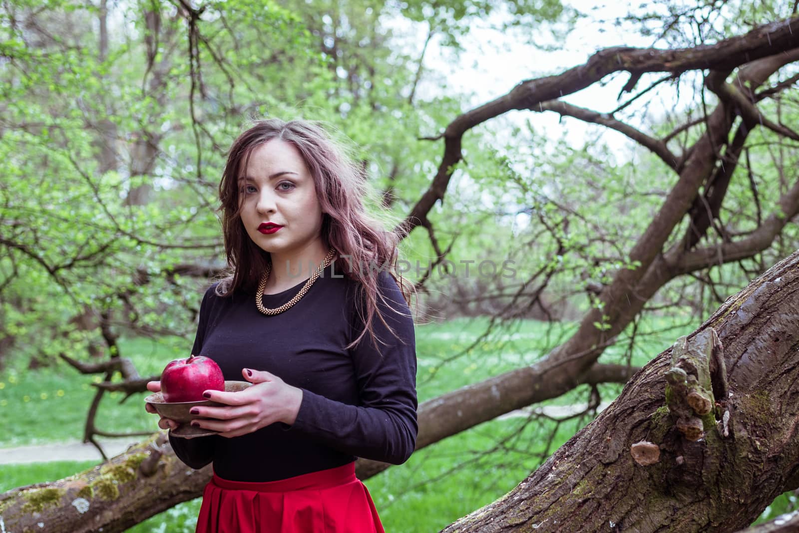 girl in a red skirt standing near a tree trunk with apple in hand