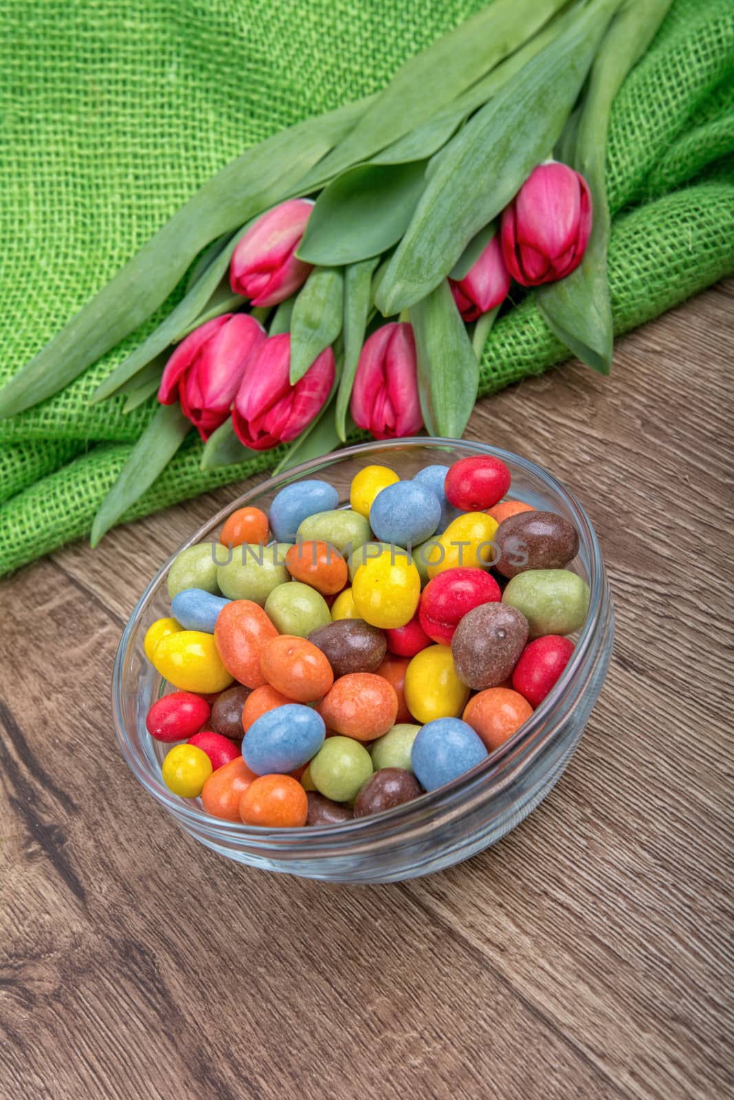 Colorful candies in a glass bowl with red tulips on a wooden background