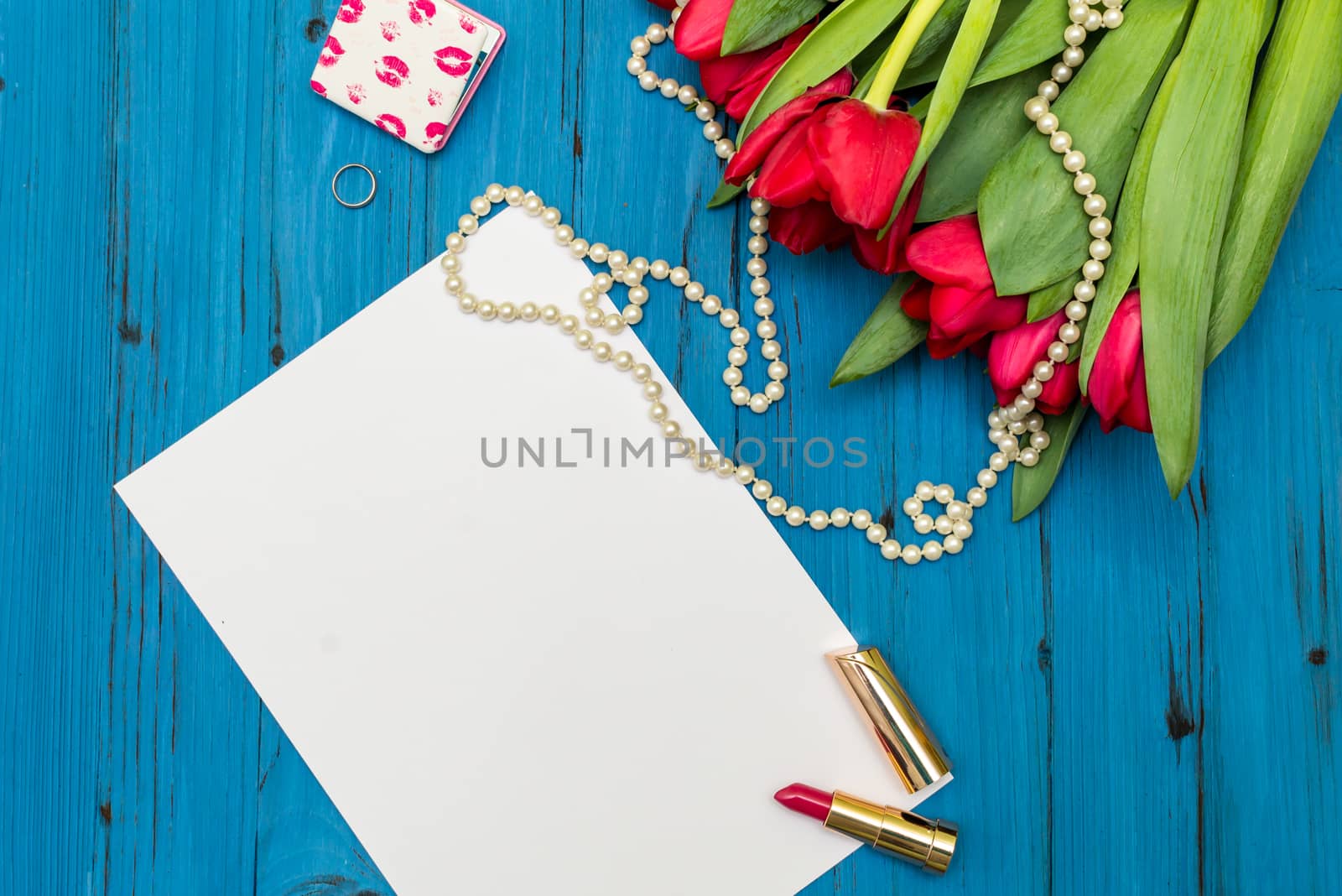 red tulips in the necklace, lipstick, one ring and a white sheet of paper on the background of blue wooden board