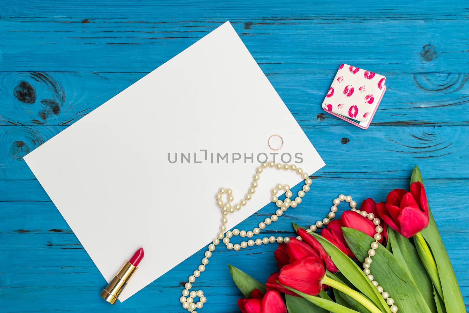 red tulips in the necklace, lipstick, one ring and a white sheet of paper on the background of blue wooden board