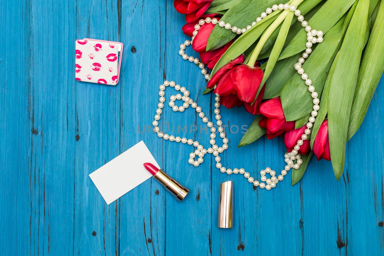 red tulips in the necklace, lipstick and a white card on the background of blue wooden board