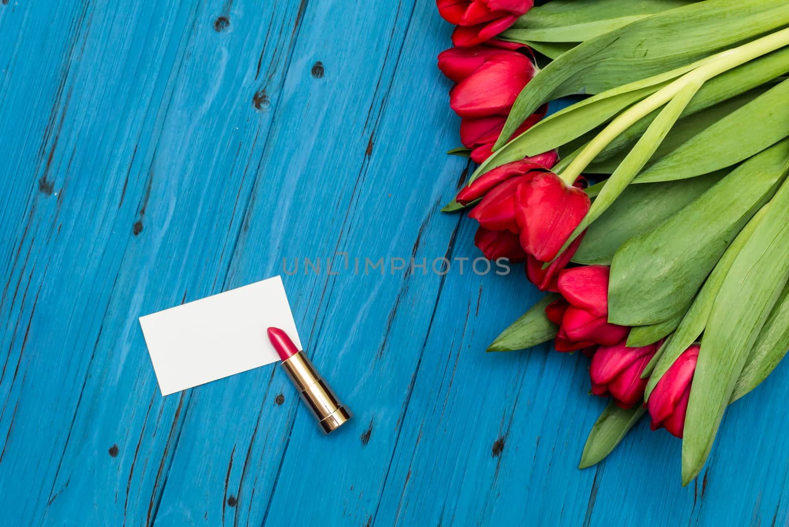 red tulips, lipstick and a white card for your greetings on the background of blue wooden board