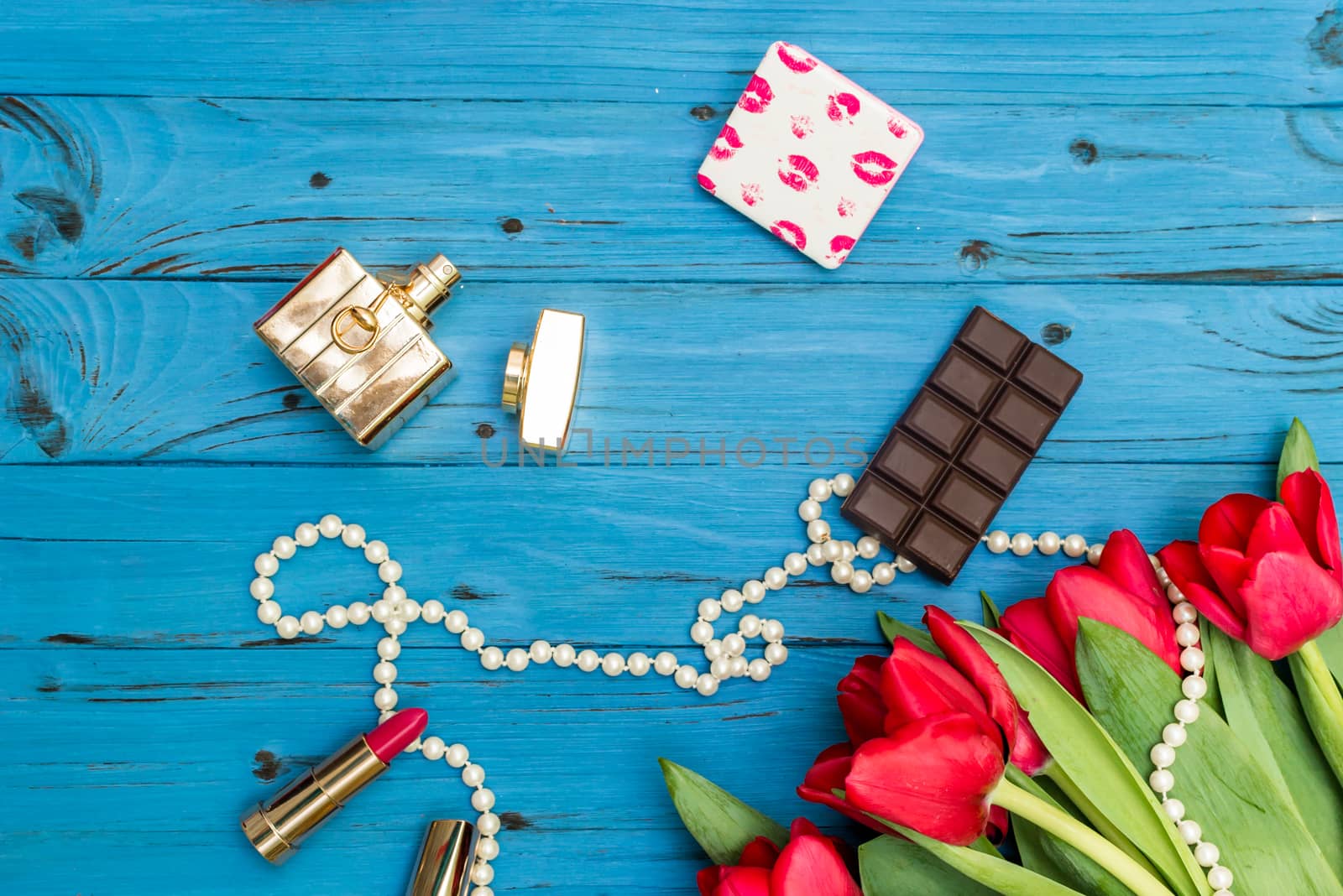 red tulips in the necklace, lipstick, bottle of perfume and piece of chocolate on the background of blue wooden board