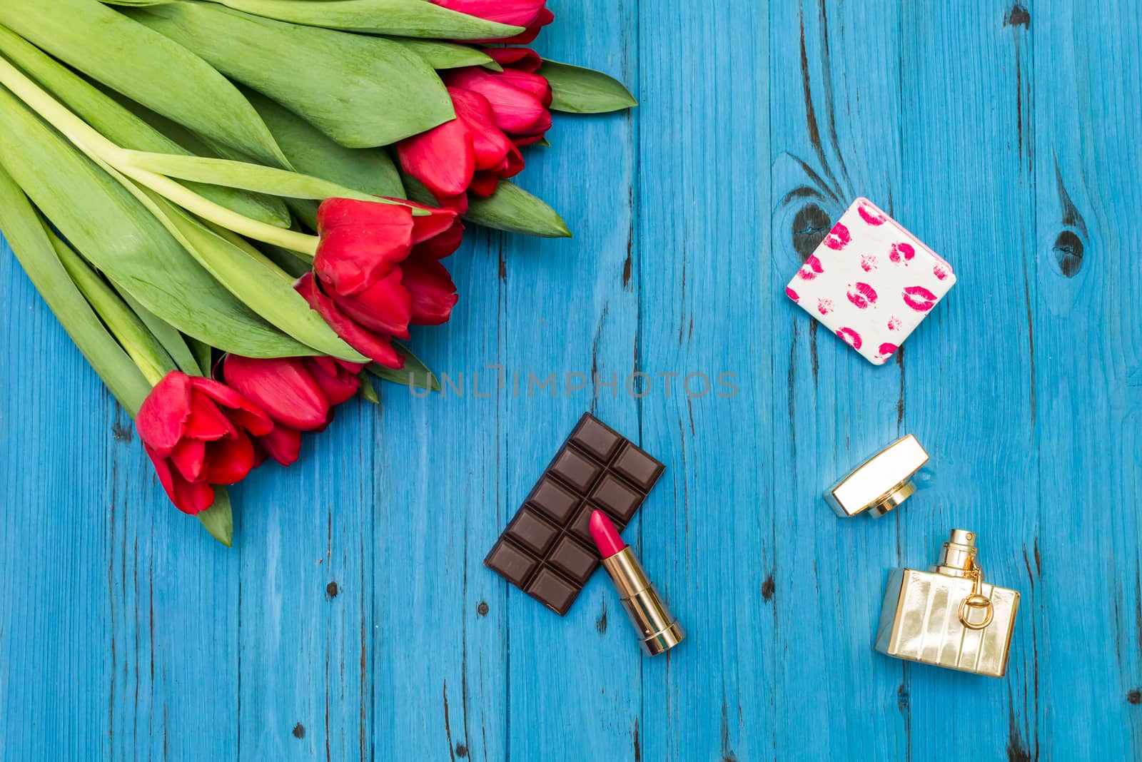 red tulips in the necklace, lipstick, bottle of perfume and piece of chocolate on the background of blue wooden board