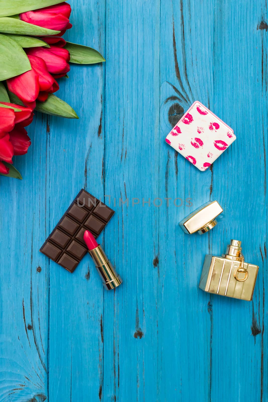 red tulips in the necklace, lipstick, bottle of perfume and piece of chocolate on the background of blue wooden board