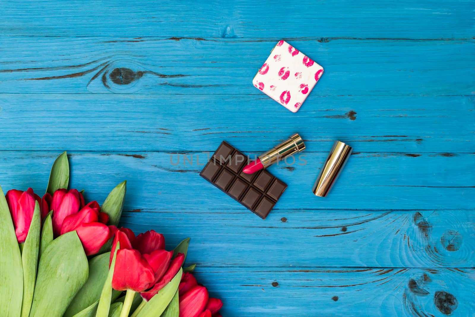 red tulips in the necklace, lipstick and piece of chocolate on the background of blue wooden board