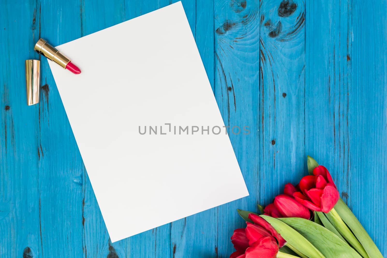 red tulips, lipstick and a white sheet of paper for your greetings on the background of blue wooden board