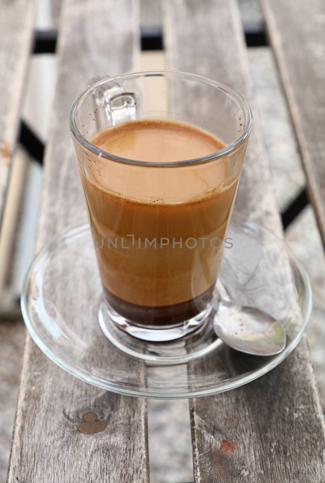 Turkish freshly brewed natural ground coffee with milk in transparent full glass cup with saucer over vintage wooden table, close up, high angle view
