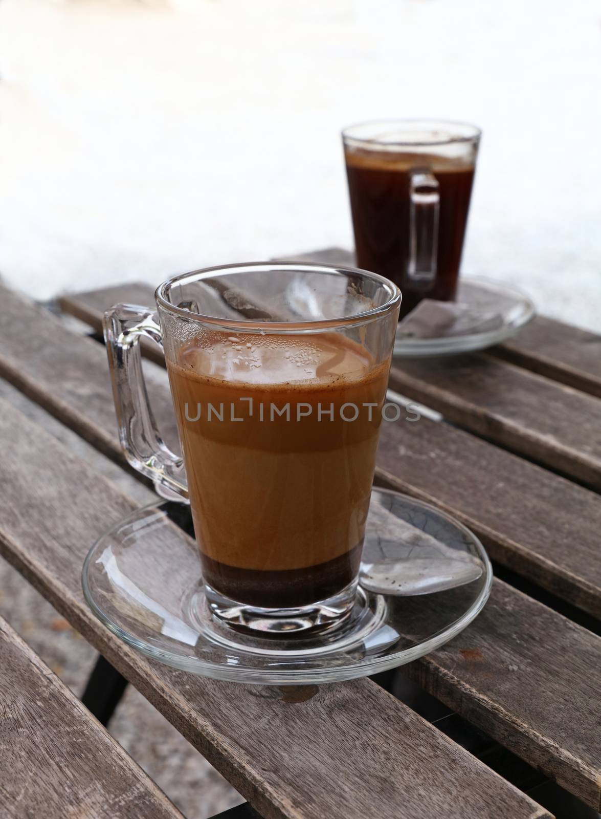 Two full Turkish freshly brewed natural ground coffee, black and with milk, in transparent full glass cups with saucer and metal spoons over vintage wooden table, close up, high angle view