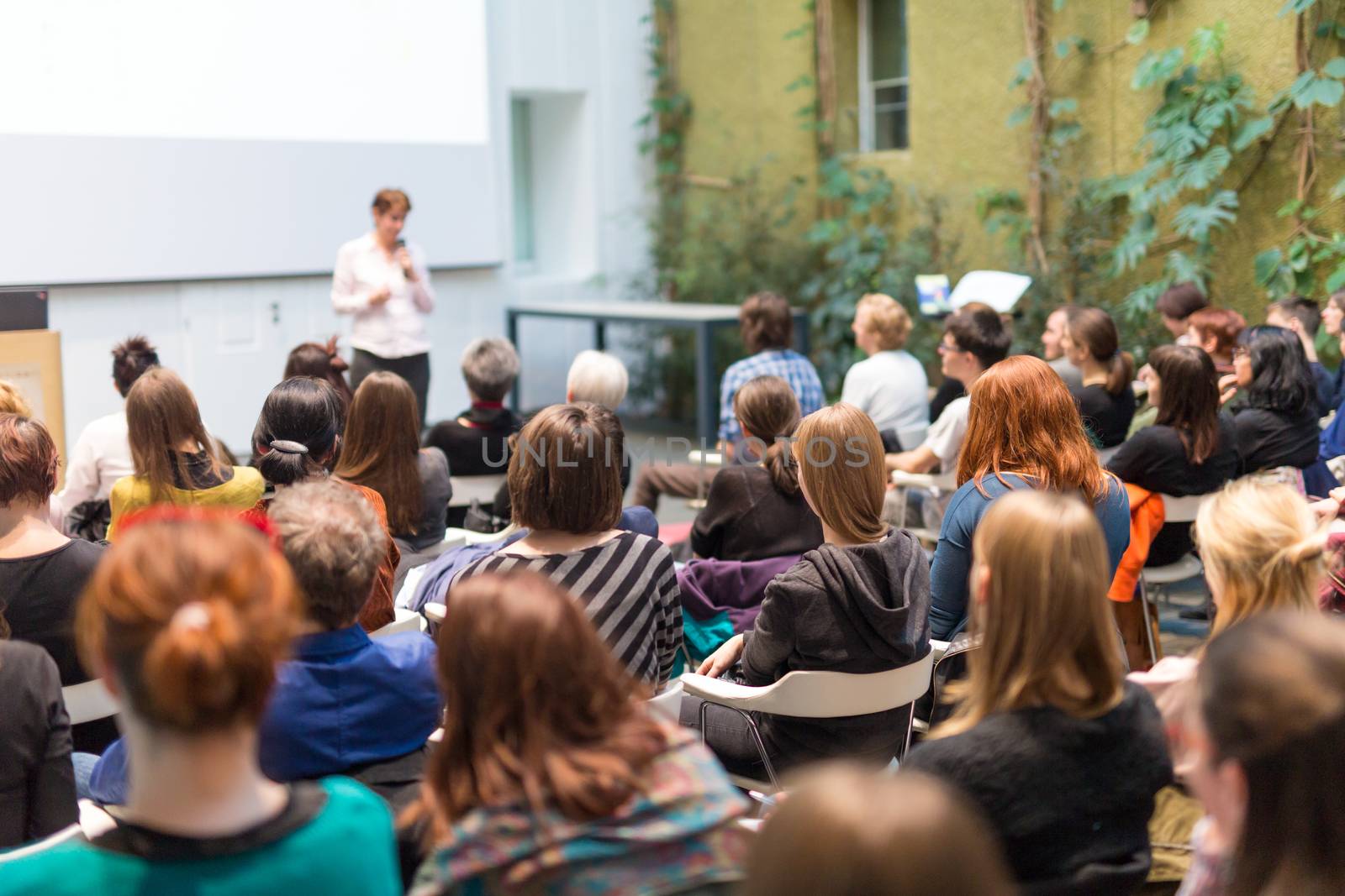 Female speaker giving presentation in lecture hall at university workshop. Audience in the conference hall. Rear view of unrecognized participant in audience. Scientific conference event.