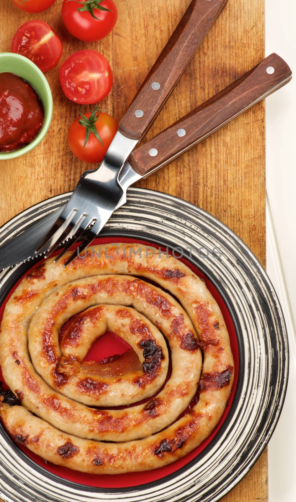 Delicious Grilled Spiral Sausage on Striped Plate with Ketchup, Cherry Tomatoes  and Rustic Fork and Knife on Wooden Cutting Board closeup on Plank background