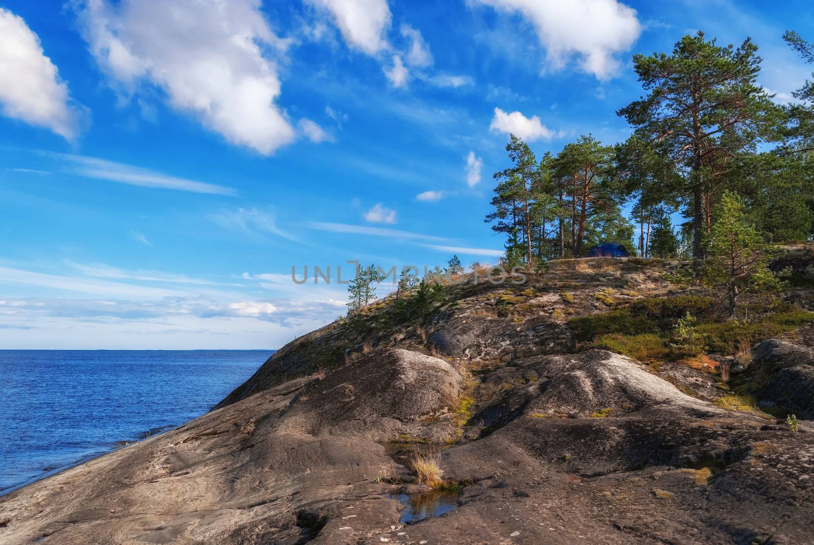 Blue tourist tent on a high rocky shore of Lake Ladoga on the background of blue sky with white clouds in Karelia.