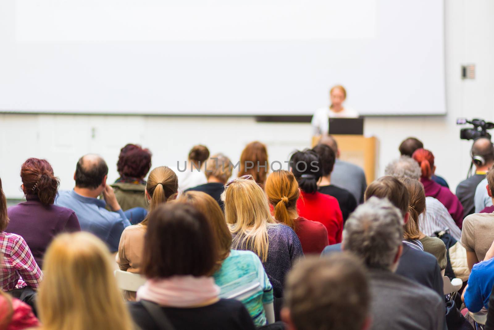 Business and entrepreneurship symposium. Female speaker giving a talk at business meeting. Audience in conference hall. Rear view of unrecognized participant in audience. Copy space on whitescreen.