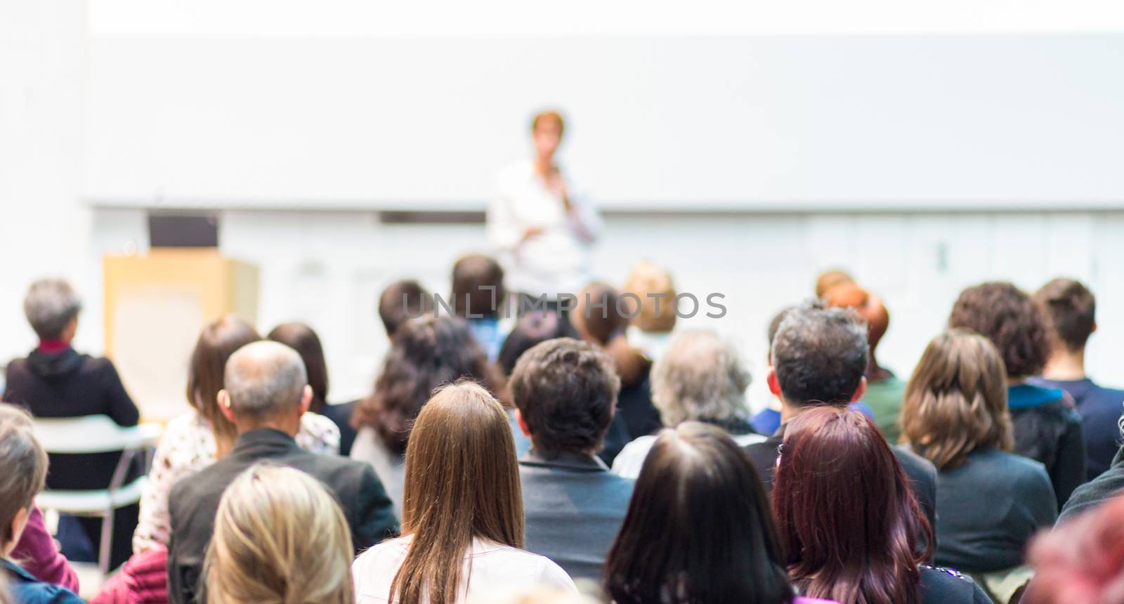 Business and entrepreneurship symposium. Female speaker giving a talk at business meeting. Audience in conference hall. Rear view of unrecognized participant in audience. Copy space on whitescreen.