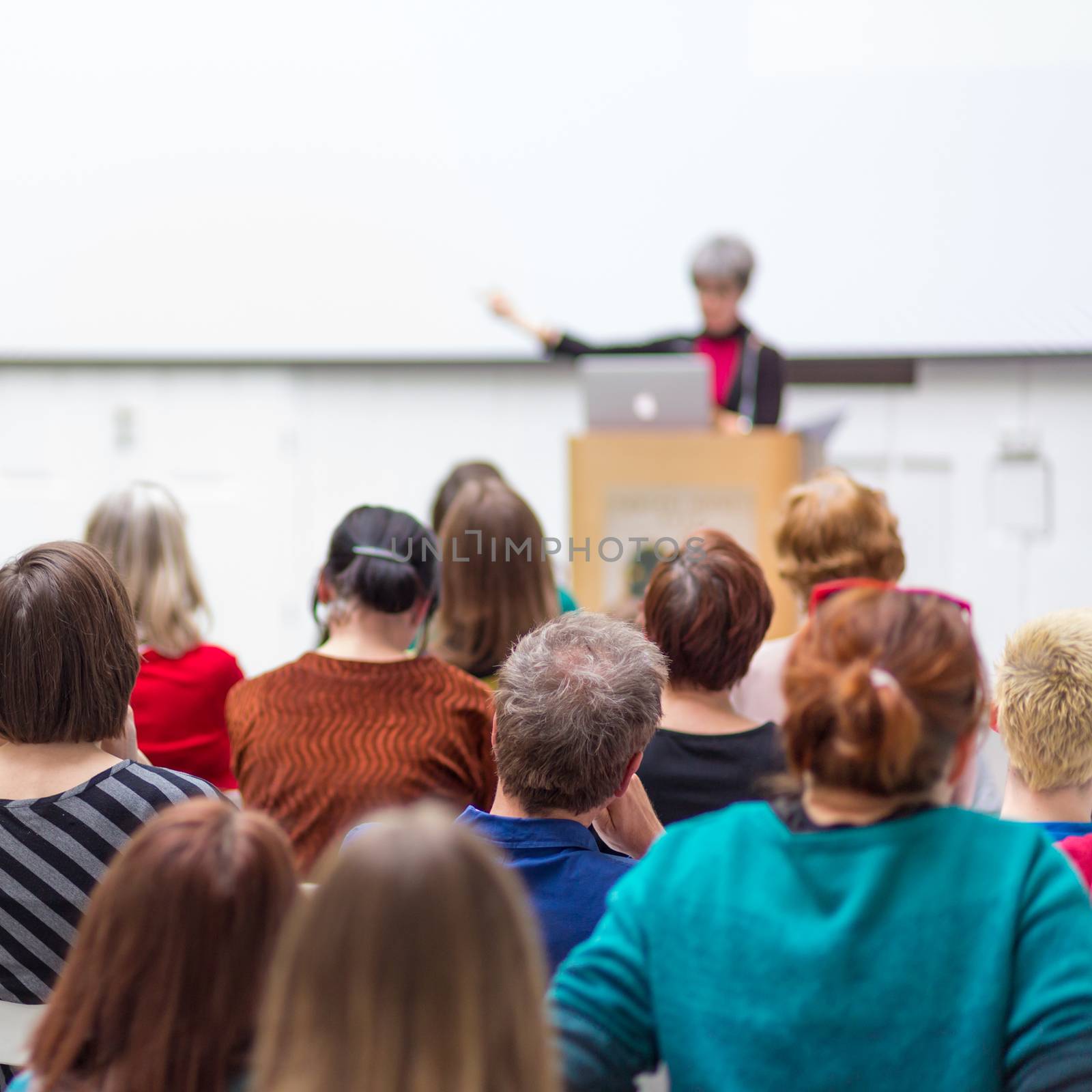 Business and entrepreneurship symposium. Female speaker giving a talk at business meeting. Audience in conference hall. Rear view of unrecognized participant in audience. Copy space on whitescreen.