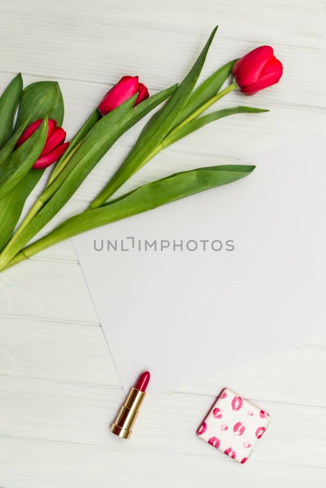 red tulips, lipstick and a white sheet of paper for your greetings on the background of white wooden board