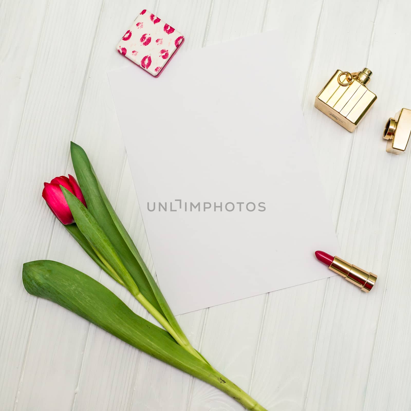one red tulip, bottle of perfume, lipstick and sheet of paper for your greetings on the background of white wooden board