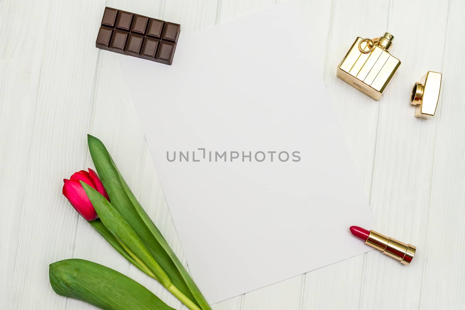 one red tulip, bottle of perfume, lipstick, piece of chocolate and sheet of paper for your greetings on the background of white wooden board