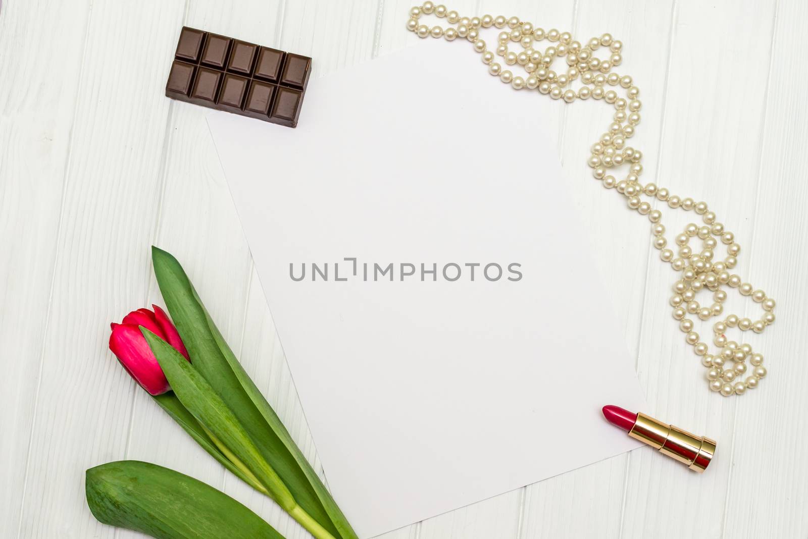 top view beautiful one red tulip, necklace, lipstick, piece of chocolate and sheet of paper for your greetings on the background of white wooden board