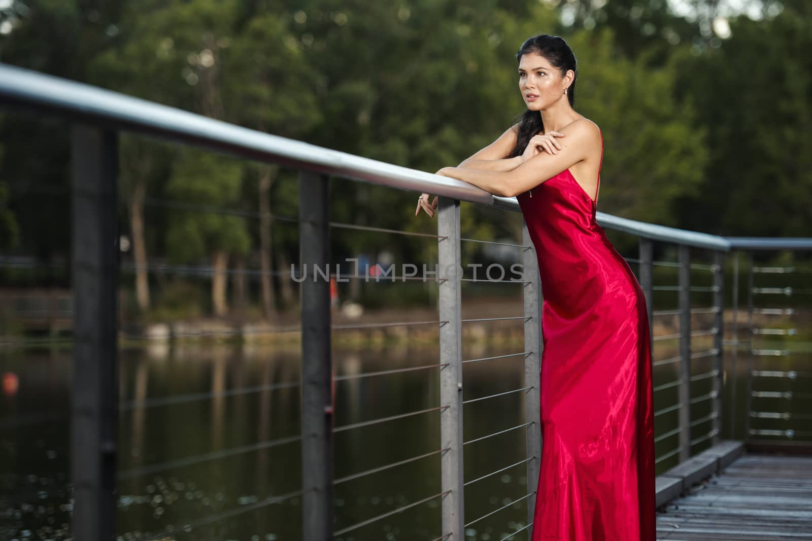 Beautiful young woman wearing a long red silk formal dress in the gardens in the afternoon. 