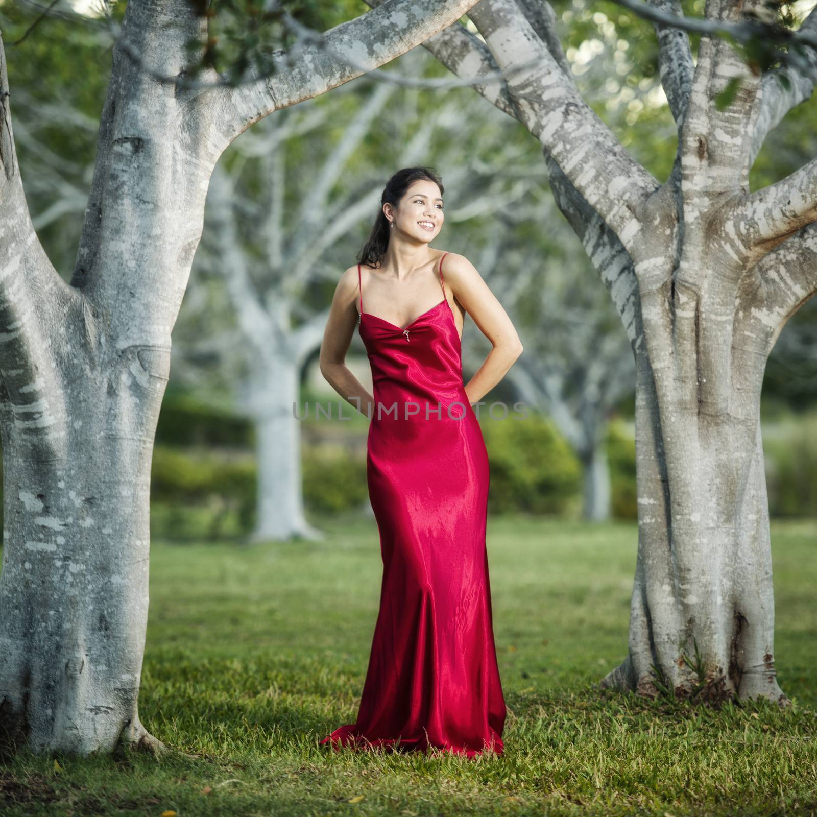 Beautiful young woman wearing a long red silk formal dress in the gardens in the afternoon. 