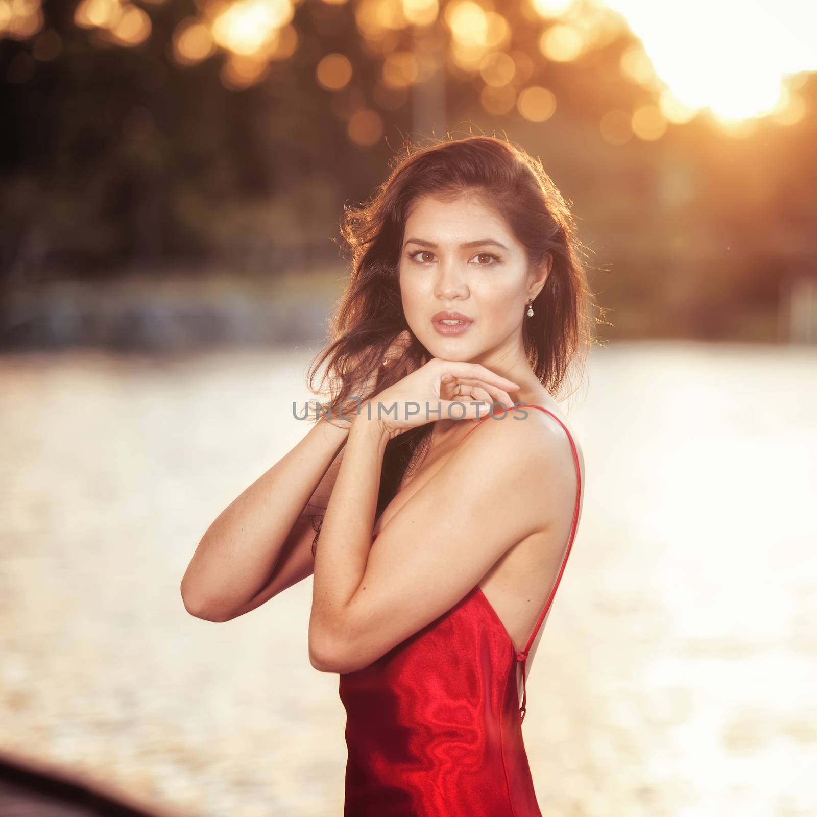 Beautiful young woman wearing a long red silk formal dress in the gardens in the afternoon. 