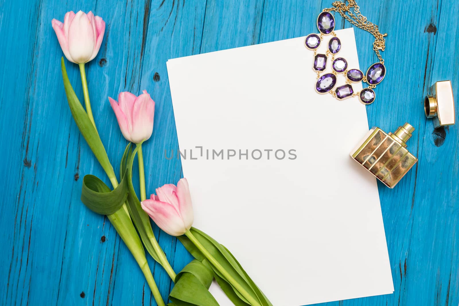 top view beautiful pink tulips, necklace, bottle of perfume and sheet of paper for your greetings on the background of blue wooden board