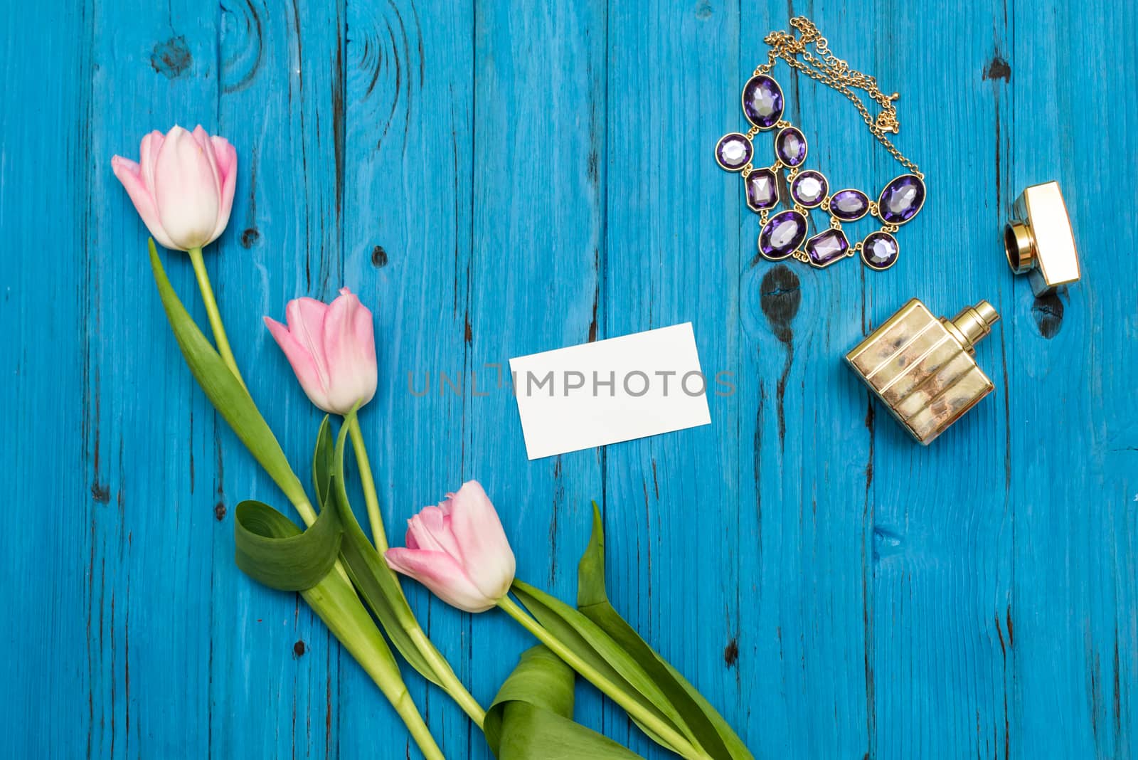 top view beautiful pink tulips, necklace, bottle of perfume and sheet of paper for your greetings on the background of blue wooden board