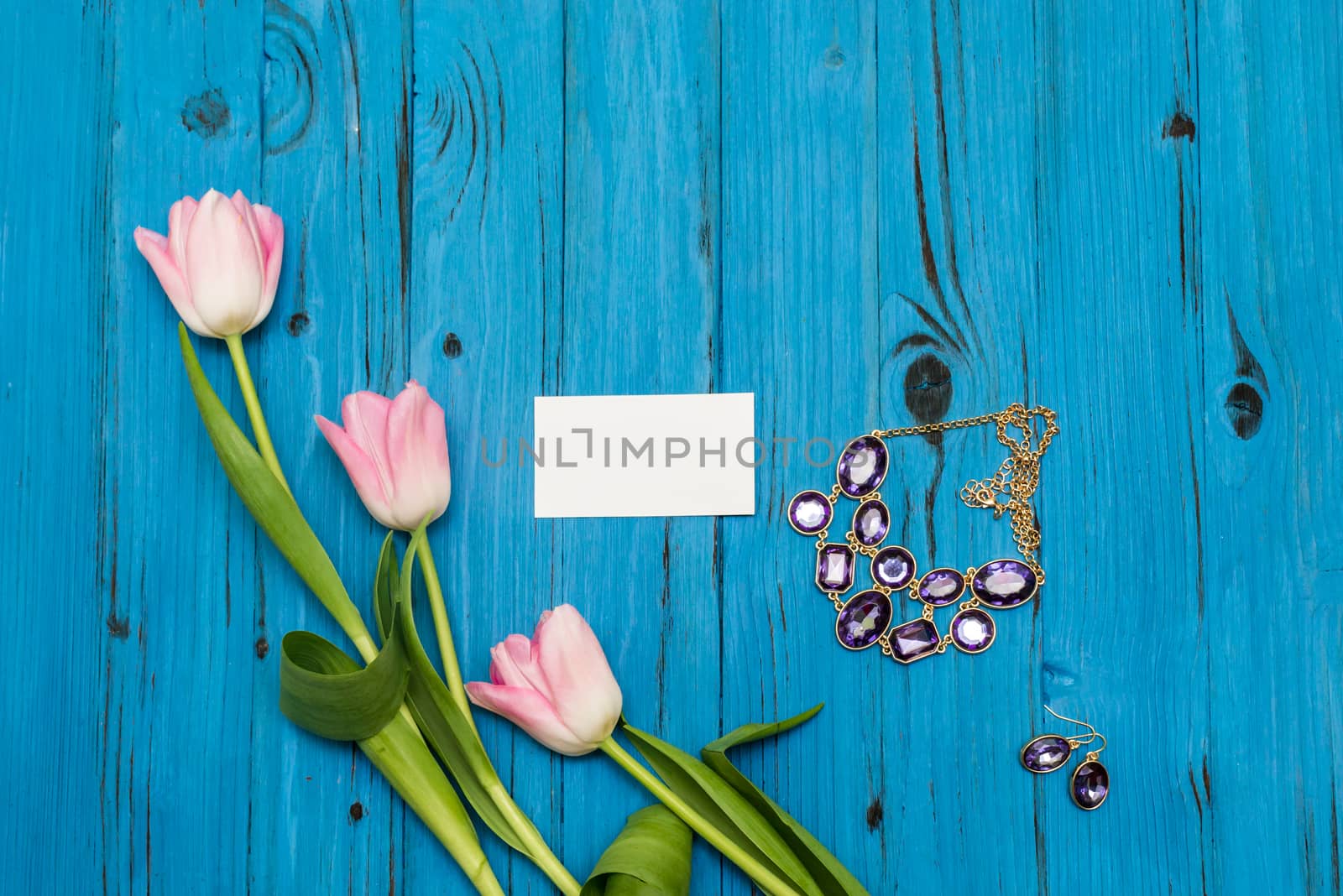 top view beautiful pink tulips, necklace with earring and card of paper for your greetings on the background of blue wooden board