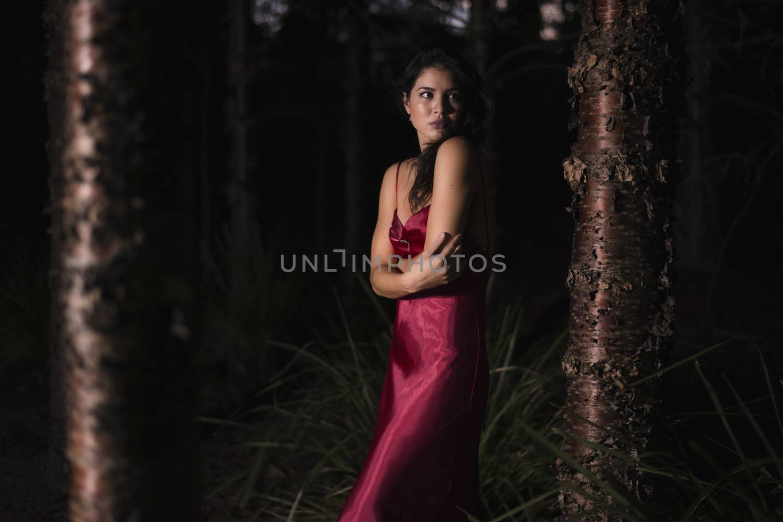 Beautiful young woman wearing a long red silk formal dress in the gardens in the early evening. 