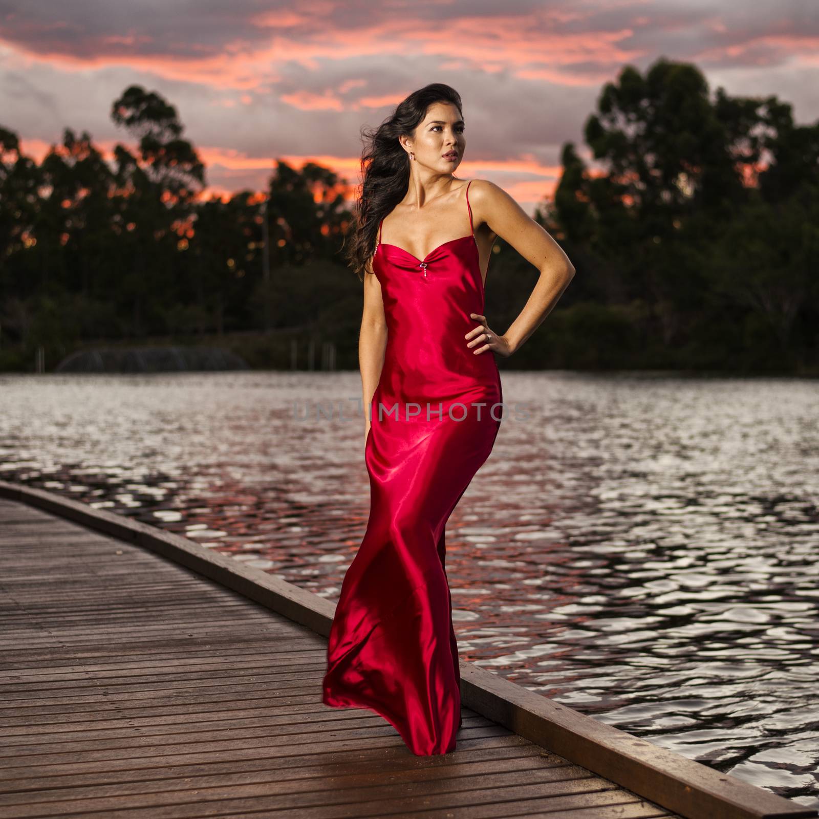 Beautiful young woman wearing a long red silk formal dress in the gardens in the afternoon.