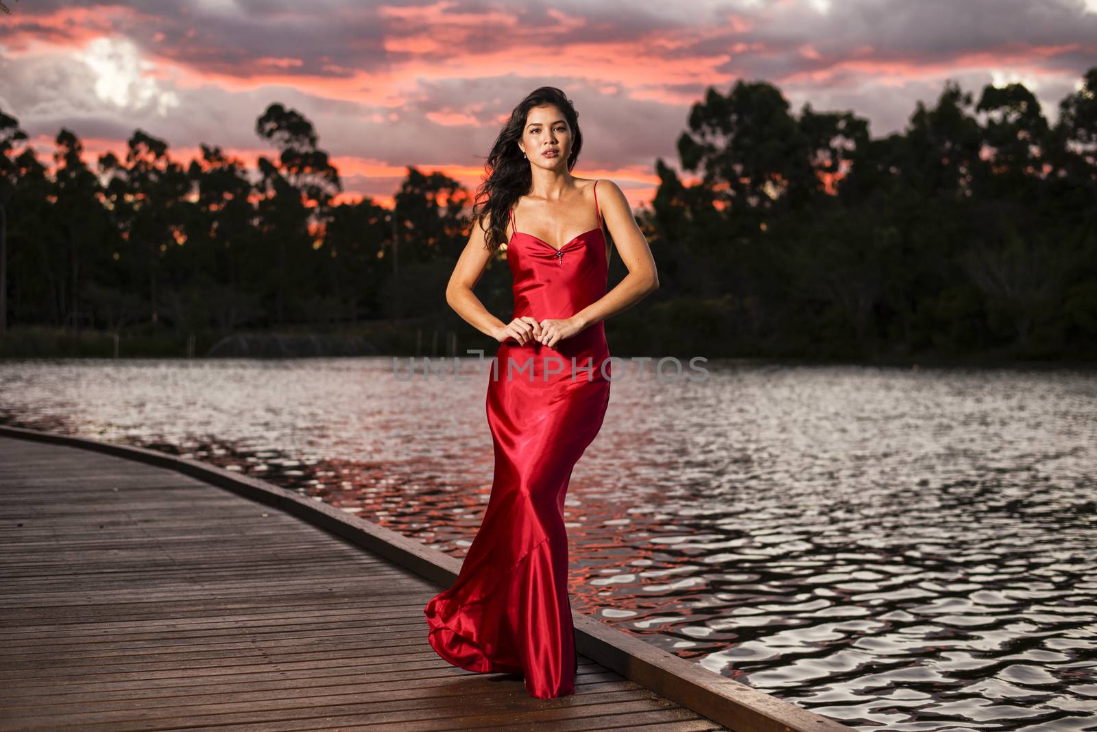 Beautiful young woman wearing a long red silk formal dress in the gardens in the afternoon.