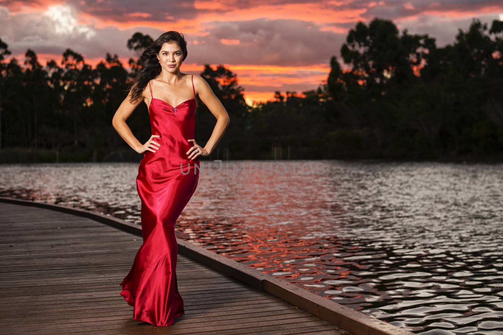 Beautiful young woman wearing a long red silk formal dress in the gardens in the afternoon. 