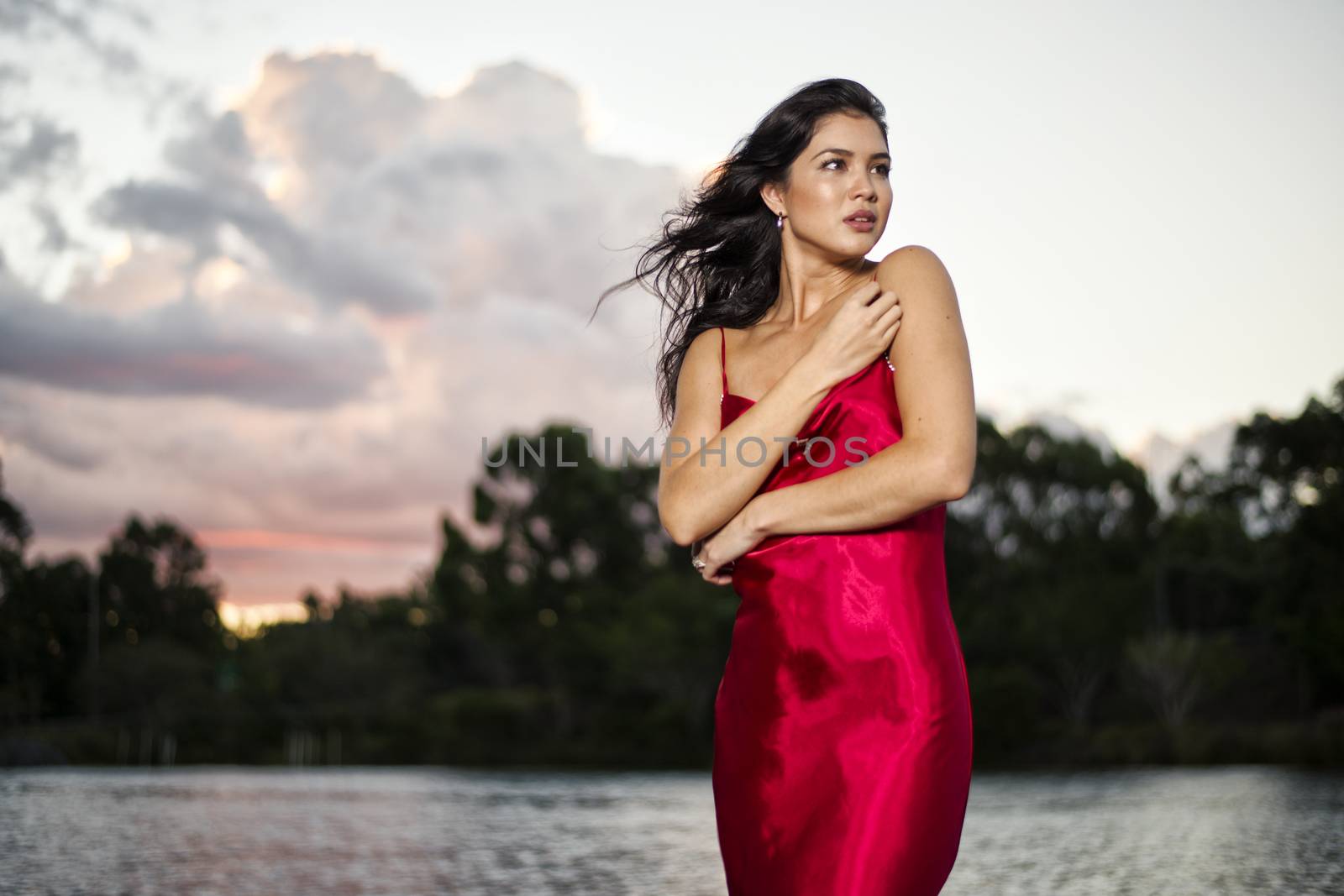 Beautiful young woman wearing a long red silk formal dress in the gardens in the afternoon.