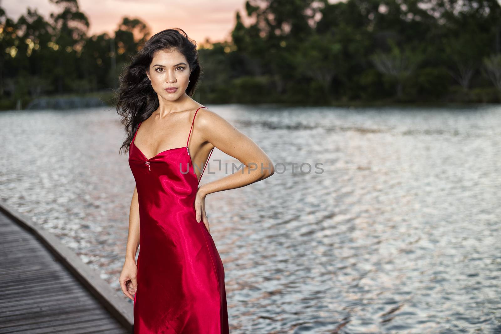 Beautiful young woman wearing a long red silk formal dress in the gardens in the afternoon. 